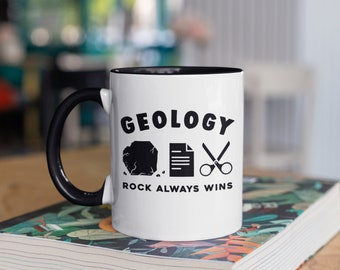 May include: White ceramic mug with a black handle and interior. The mug features the word "GEOLOGY" above a rock, paper, and scissors graphic, with "ROCK ALWAYS WINS" below. The mug is sitting on a stack of books.