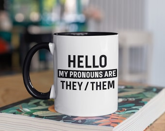 May include: White ceramic mug with a black handle and interior. The mug features the text "HELLO MY PRONOUNS ARE THEY / THEM" in black, centered on the front. The mug is sitting on a book with a colorful floral design.
