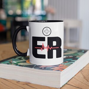 May include: White ceramic mug with a black handle and interior. The mug features the letters "ER" in bold black font, a red heartbeat line, and a white cross symbol. The mug is sitting on a book with a floral design.
