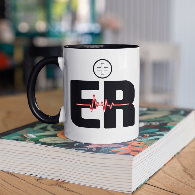 May include: White ceramic mug with a black handle and interior. The mug features the letters "ER" in bold black font, a red heartbeat line, and a white cross symbol. The mug is sitting on a book with a floral design.