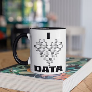 Puede incluir: Taza de cerámica blanca con interior y asa negros. La taza presenta una forma de corazón hecha de código binario, con las palabras "I DATA" impresas en negro. La taza está sobre un libro con un diseño floral.