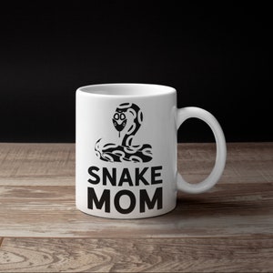 Pode incluir: Caneca de cerâmica branca com um desenho preto de cobra e a frase "SNAKE MOM". O desenho da cobra é estilizado com padrões intrincados. A caneca tem uma alça curva. Está colocada sobre uma superfície de madeira e um fundo escuro.