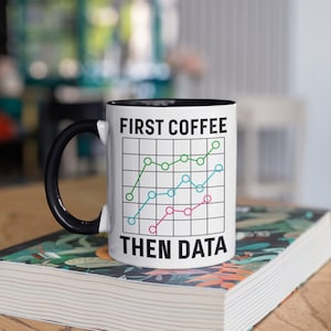 May include: White ceramic mug with a black handle and interior. The mug features the text "FIRST COFFEE THEN DATA" above a graph with green, blue, and pink lines. The mug is sitting on a book with a floral design.