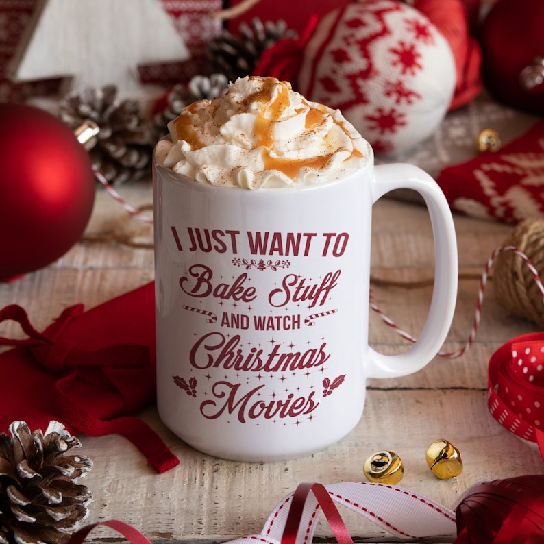 May include: A white ceramic mug filled with whipped cream and caramel, with the text "I JUST WANT TO Bake Stuff AND WATCH Christmas Movies" in red. The mug is surrounded by Christmas decorations, including ornaments and ribbons.