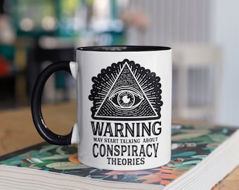 Peut inclure: Mug en céramique blanche avec une anse et un intérieur noirs. Le mug présente un graphisme noir d'un œil dans un triangle, avec le texte "WARNING MAY START TALKING ABOUT CONSPIRACY THEORIES". Le mug est posé sur un livre.