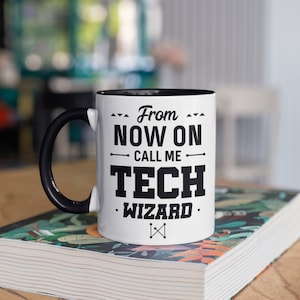 May include: White ceramic mug with a black handle and interior. The mug features the text "From Now On Call Me Tech Wizard" in bold black letters. The mug is sitting on a book with a floral design.