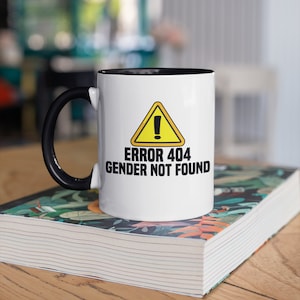 Könnte beinhalten: Weißer Keramikbecher mit schwarzem Griff und Innenrand. Der Becher zeigt den Text "ERROR 404 GENDER NOT FOUND" in schwarzer Schrift, mit einem gelben Warnschild. Der Becher steht auf einem Buch mit floralem Design.