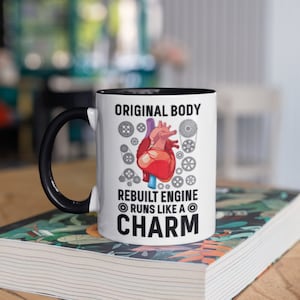 Könnte beinhalten: Weißer Keramikbecher mit schwarzem Griff und Innenraum. Der Becher zeigt eine Grafik eines menschlichen Herzens, Zahnräder und den Text "ORIGINAL BODY REBUILT ENGINE RUNS LIKE A CHARM."