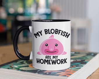 May include: White ceramic mug with a black handle and interior. The mug features a pink blobfish illustration and the text "MY BLOBFISH ATE MY HOMEWORK" in black. The mug is on a book with a colourful floral design.