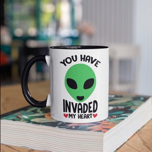 May include: White ceramic mug with a black handle and interior. The mug features a green alien face design with the text "YOU HAVE INVADED MY HEART" in black. Two red hearts are below the text.