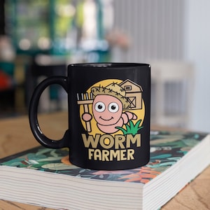 May include: Black ceramic mug featuring a cartoon worm character wearing a straw hat and holding a rake. The mug has the words "WORM FARMER" in gold lettering, set against a yellow circle with a barn illustration.