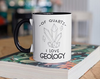 May include: White ceramic mug with a black handle and interior. The mug features a black and white graphic of quartz crystals and the text "-OF QUARTZ- I LOVE GEOLOGY". The mug is sitting on a book with a floral design.