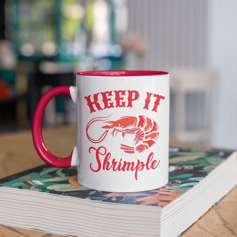 May include: White ceramic mug with a red rim and handle. The mug features a red graphic of a shrimp with the text "Keep it Shrimple" printed on it.