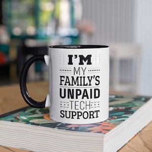 Puede incluir: Taza de café blanca con borde y asa negros. La taza tiene texto negro que dice "Soy el soporte técnico no remunerado de mi familia."