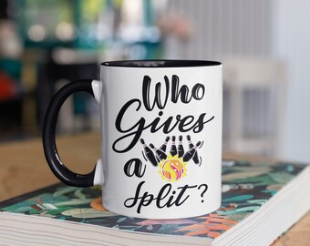 Pode incluir: Caneca de cerâmica branca com alça e interior pretos. A caneca apresenta o texto "Who Gives a Split?" em escrita preta, juntamente com um gráfico de pinos de boliche e uma bola. A caneca está em um livro.