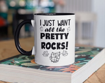 May include: White ceramic mug with a black handle and interior, featuring the text "I JUST WANT all the PRETTY ROCKS!" in bold black letters. The mug is decorated with crystal illustrations and sits on a book with a floral cover.