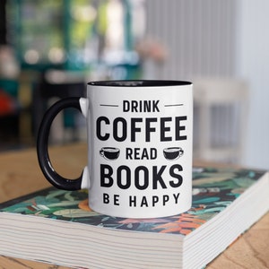 May include: White ceramic mug with a black handle and interior, featuring the text "DRINK COFFEE READ BOOKS BE HAPPY" in black. The mug is sitting on top of a book with a floral design.