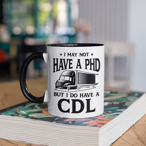 May include: White ceramic coffee mug with a black rim and handle. The mug has a black and white graphic of a semi-truck with the text "I may not have a PHD but I do have a CDL".