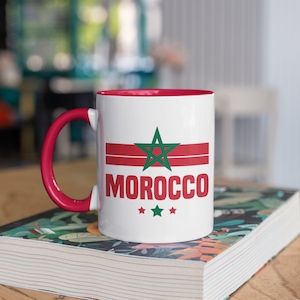 May include: White ceramic mug with a red interior and handle. The mug features the word "MOROCCO" in red, a green star, and red stripes, with three green stars below. The mug is sitting on a book.