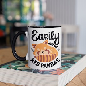 Peut inclure: Mug en c&eacute;ramique blanche avec une anse et un int&eacute;rieur noirs. Le mug affiche le texte "Easily distracted by Red Pandas" avec une illustration de panda roux. Le mug est pos&eacute; sur un livre &agrave; motifs floraux.