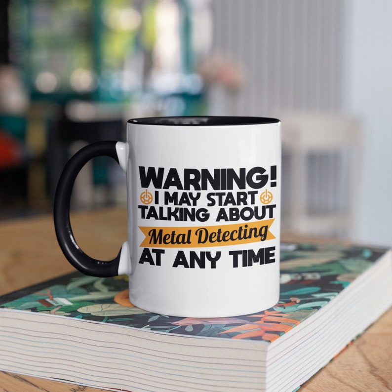 May include: White ceramic mug with a black interior and handle. The mug features the text "WARNING! I MAY START TALKING ABOUT Metal Detecting AT ANY TIME" in black and orange lettering. The mug is sitting on a book.