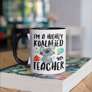 May include: White ceramic mug with a black handle and black rim. The mug features a cartoon koala wearing glasses and holding an apple. The text "I'm a highly koalafied teacher" is printed on the mug.