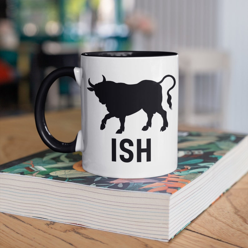 K&ouml;nnte beinhalten: Wei&szlig;e Keramiktasse mit schwarzem Rand und Henkel. Die Tasse zeigt eine schwarze Silhouette eines Stiers und das Wort "ISH" in schwarzen Buchstaben.