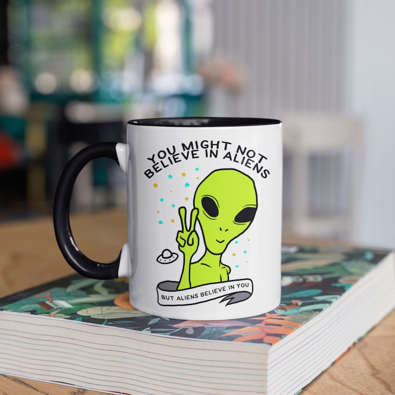 Puede incluir: Taza de cer&aacute;mica blanca con asa e interior negros. La taza presenta un gr&aacute;fico de un alien&iacute;gena verde haciendo el signo de la paz, con el texto "YOU MIGHT NOT BELIEVE IN ALIENS BUT ALIENS BELIEVE IN YOU".