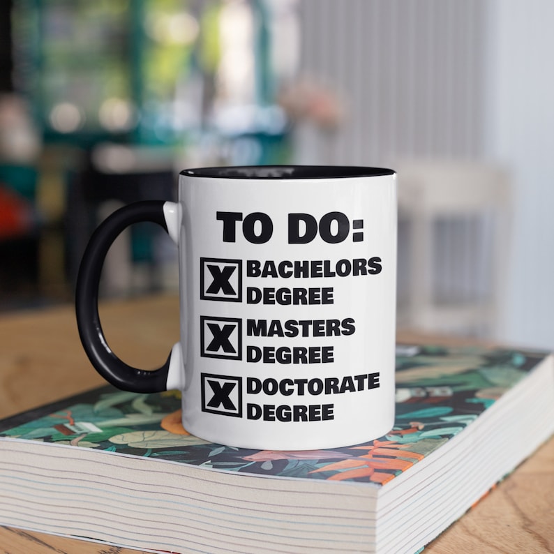 Puede incluir: Taza de cer&aacute;mica blanca con asa e interior negros. La taza presenta el texto "TO DO:" seguido de casillas de verificaci&oacute;n para los t&iacute;tulos de Licenciatura, M&aacute;ster y Doctorado. La taza est&aacute; sobre un libro con un dise&ntilde;o floral colorido.