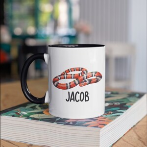 May include: White ceramic mug with a black handle and interior. The mug features a colorful illustration of a snake with red, black, and white bands, and the name "JACOB" in black text. The mug is sitting on a book.