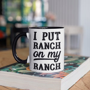 May include: White ceramic mug with a black rim and handle. The mug has black text that reads "I PUT RANCH on my RANCH".
