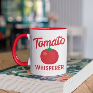 May include: White ceramic mug with a red rim and handle. The mug has a red tomato graphic and the text "Tomato Whisperer" printed on it.