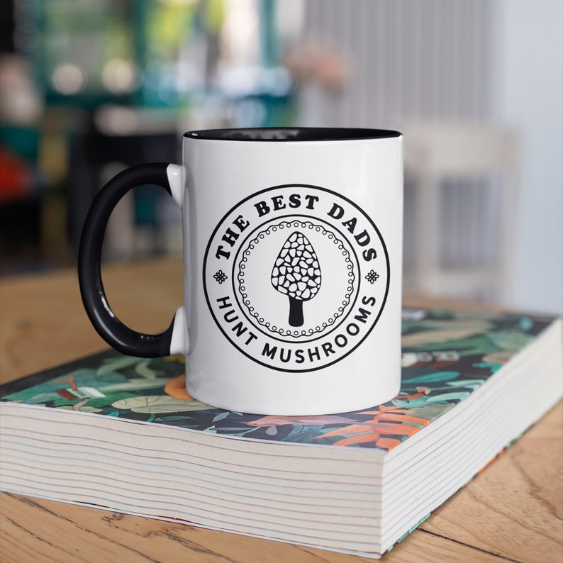 May include: White ceramic mug with a black handle and interior. The mug features a circular design with the text "THE BEST DADS HUNT MUSHROOMS" around a morel mushroom illustration. The mug is resting on a book with a floral cover.