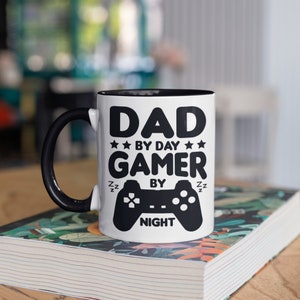 May include: White ceramic mug with a black interior and handle. The mug features the text "DAD BY DAY GAMER BY NIGHT" in black, along with a controller graphic. The mug is sitting on a book with a floral design.