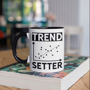 May include: White ceramic mug with a black interior and handle. The mug features the words "TREND SETTER" in bold black letters, along with a black and white line graph design. The mug is sitting on a book.