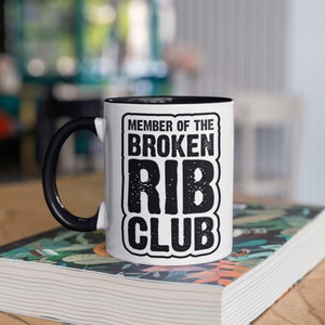 Puede incluir: Taza de cerámica blanca con asa e interior negros. La taza presenta el texto "MEMBER OF THE BROKEN RIB CLUB" en una fuente audaz y desgastada. La taza está sobre un libro con un diseño floral colorido.