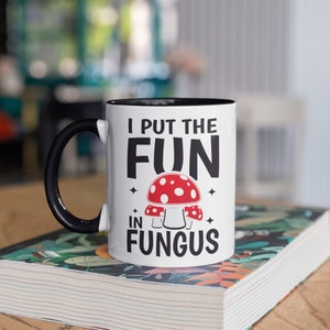 May include: White ceramic mug with a black handle and interior. The mug features the text "I PUT THE FUN IN FUNGUS" with a red and white mushroom graphic. The mug is sitting on a book with a floral design.