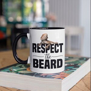 May include: White ceramic mug with a black rim and handle. The mug features a bearded dragon lizard with the text "Respect the Beard" in black.