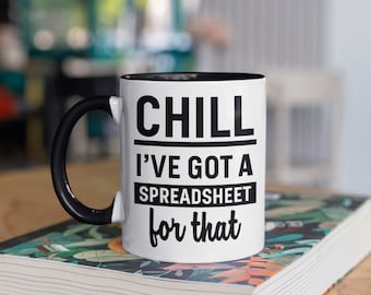 May include: White ceramic mug with a black handle and interior. The mug features the text "CHILL I'VE GOT A SPREADSHEET for that" in black, centered on the front. The mug is resting on a book with a floral design.