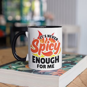 May include: White ceramic mug with a black handle and interior. The mug features the text "IT'S NEVER SPICY ENOUGH FOR ME" in red, orange, and black, with a chili pepper graphic. The mug is sitting on a book with a floral design.