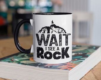 May include: White ceramic mug with a black handle and interior. The mug features the text "WAIT I SEE A ROCK" in black, with a mountain silhouette graphic. The mug is sitting on a book with a floral design.