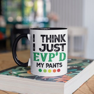 May include: White coffee mug with a black handle and black rim. The mug has a green and black graphic that says "I think I just EVP'd my pants" with a row of five green, yellow, orange, and red dots below the text.