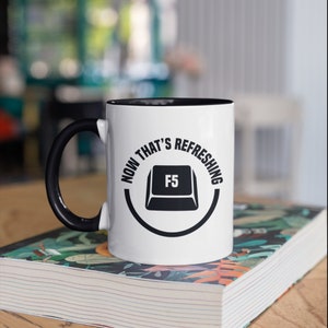Puede incluir: Taza de cerámica blanca con interior y asa negros. La taza presenta el texto "NOW THAT'S REFRESHING" alrededor de un gráfico de la tecla F5. La taza está sobre un libro.