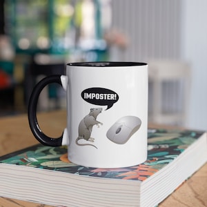 Może przedstawiać: Biały ceramiczny kubek z czarnym uchwytem i wnętrzem. Kubek przedstawia kreskówkowego szczura z dymkiem z napisem "IMPOSTER!" i myszką komputerową. Kubek stoi na książce z kolorowym kwiatowym wzorem.