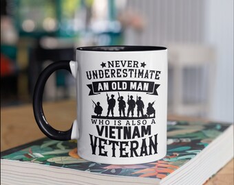 May include: White ceramic mug with a black handle and interior. The mug features the text "NEVER UNDERESTIMATE AN OLD MAN WHO IS ALSO A VIETNAM VETERAN" in black, along with silhouettes of soldiers.