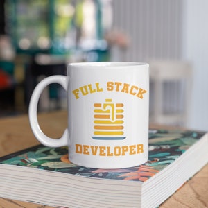 Op de afbeelding: Witte keramische mok met de tekst "FULL STACK DEVELOPER" in oranje en een afbeelding van een stapel pannenkoeken. De mok staat op een boek met een bloemenpatroon.