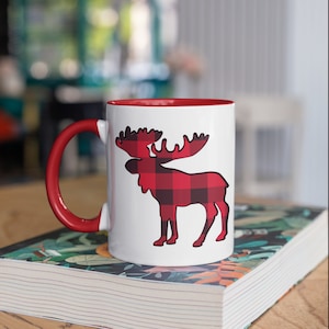 May include: White ceramic mug with a red interior and handle. The mug features a black outline of a moose filled with a red and black buffalo plaid pattern. The mug is sitting on a book.
