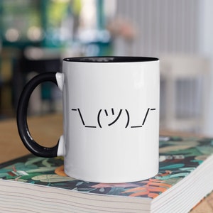 May include: White ceramic mug with a black interior and handle. The mug features a black shrug emoticon on the front. The mug is sitting on a stack of books.