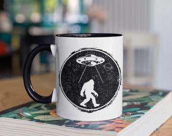 Può includere: Una tazza da caffè bianca con un bordo e un manico neri. La tazza presenta una grafica in bianco e nero di un Bigfoot che cammina sotto un UFO.