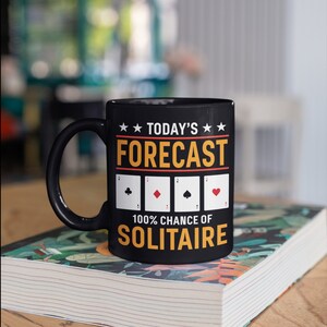 May include: Black coffee mug with a white design that says "Today's Forecast 100% Chance of Solitaire" with four playing card symbols.